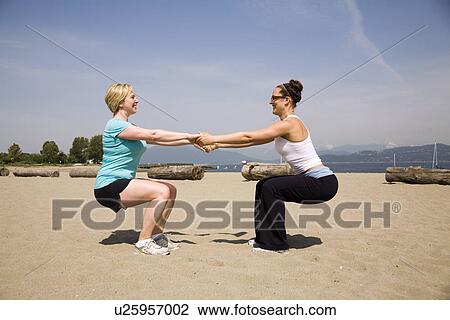 Stock Image - Two women holding hands doing squats exercising at beach, Vancouver, BC. Fotosearch