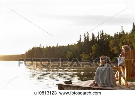 Stock Image - Two women seated on dock at sunset, Clear Lake, Manitoba, Canada. Fotosearch