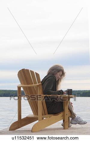 Stock Image - Woman dialing on cellphone, seated in deck chair on dock, Clear Lake, Manitoba, Canada. Fotosearch