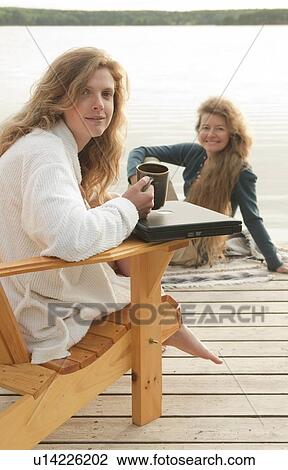 Stock Image - Woman in deck chair smiling at camera, second woman seated on dock, Clear Lake, Manitoba, Canada. Fotosearch