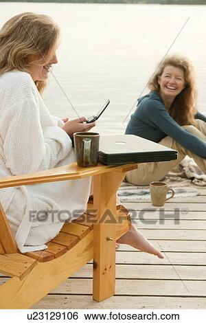 Stock Photograph - Woman in deck chair using cell phone, second woman seated on dock, Clear Lake, Manitoba, Canada. Fotosearch