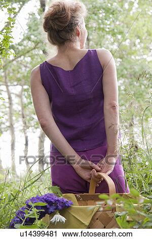 Stock Image - Woman in mid-50's, back to camera, with basket of flowers in grove of trees, Manitoba Canada. Fotosearch