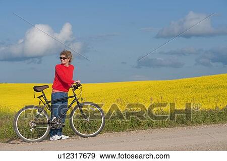 Woman in mid-50's with bike on rural road beside blooming field of canola, Manitoba, Canada View Large Photo Image Stock Photo - Woman in mid-50's with bike on rural road beside blooming field of canola, Manitoba, Canada. Fotosearch