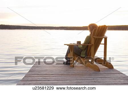 Stock Photo - Woman seated in deck chair on dock at sunset, Clear Lake, Manitoba. Fotosearch