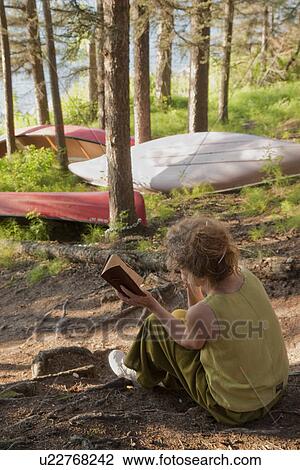 Stock Image - Woman seated in grove of trees beside lake reading a book, Clear Lake, Riding Mountain National Park, Manitoba, Canada. Fotosearch