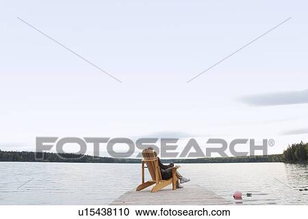 Stock Image - Woman seated on deck chair on dock, Clear Lake, Manitoba, Canada. Fotosearch