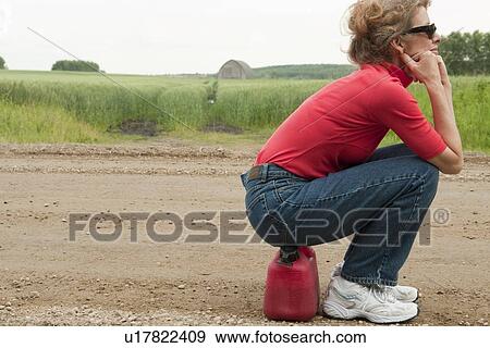 Stock Photo - Woman seated on jerry can, gravelled rural road, Manitoba, Canada. Fotosearch