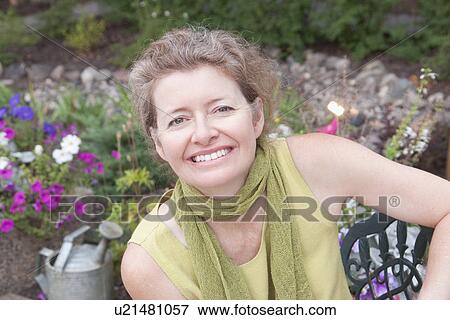Stock Photo - Woman seated on metal seat in flower garden, Manitoba, Canada. Fotosearch