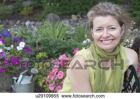 Stock Photography - Woman seated on metal seat in flower garden, Manitoba, Canada. Fotosearch