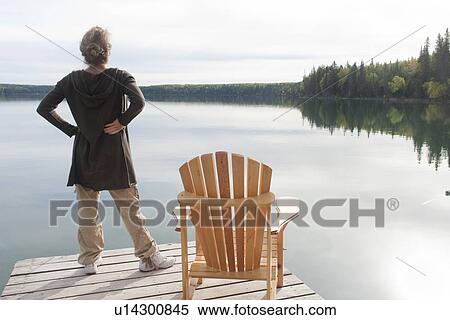 Stock Photography - Woman standing on dock beside deck chair, Clear Lake, Manitoba, Canada. Fotosearch