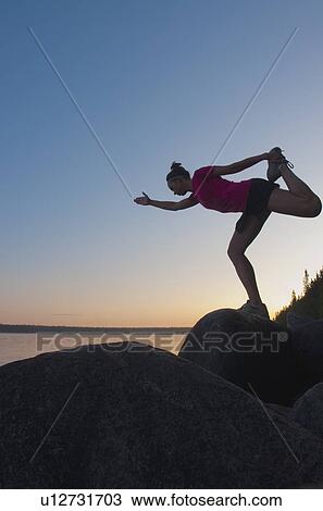 Stock Image - Woman stretching, on rock by shore, sunset, Clear Lake, Riding Mountain National Park, Manitoba, Canada. Fotosearch