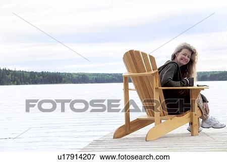 Stock Photo - Woman with cup seated on deck chair on dock, Clear Lake, Manitoba, Canada. Fotosearch