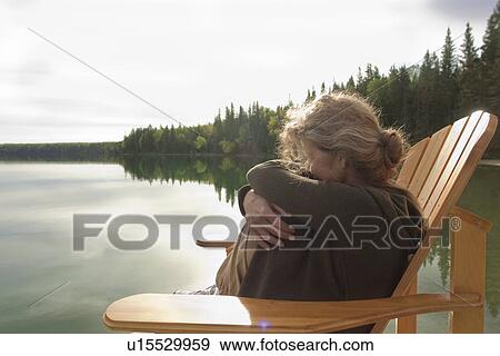 Stock Photo - Woman with knees pulled up to her chest seated in deck chair on dock, Clear Lake, Manaitoba, Canada. Fotosearch