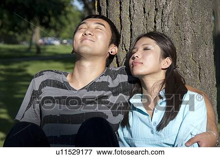 Young couple seated at base of tree, faces lifted upard. View Large Photo Image Stock Photo - Young couple seated at base of tree, faces lifted upard.. Fotosearch