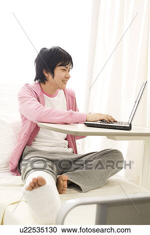 Stock Image - Man with broken leg using his laptop in the hospital. Fotosearch