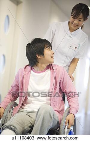 Stock Image - Nurse pushing patient in wheelchair along a corridor. Fotosearch