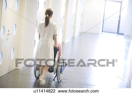 Stock Image - Nurse pushing patient in wheelchair along a corridor. Fotosearch