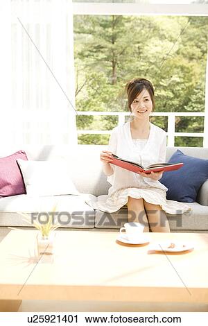 Stock Image - Woman reading book on sofa in living room. Fotosearch