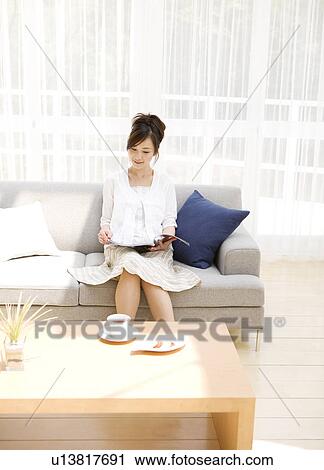 Stock Image - Woman reading magazine on sofa in living room. Fotosearch