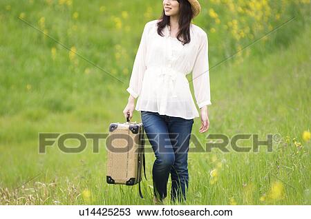 Stock Image - Woman walking through field carrying suitcase. Fotosearch