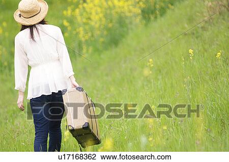Stock Image - Woman walking through field carrying suitcase. Fotosearch