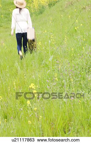 Stock Image - Woman walking through field carrying suitcase. Fotosearch