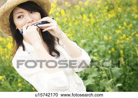 Stock Image - Woman wearing straw hat playing harmonica in a meadow. Fotosearch
