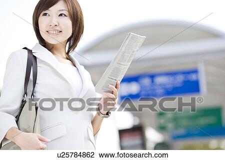 Stock Image - Businesswoman holding newspaper at bus stop. Fotosearch