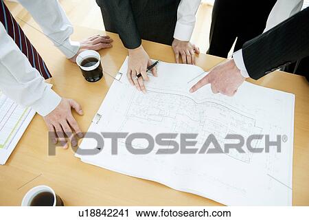 Stock Image - Colleagues working at desk in office with blueprints. Fotosearch