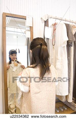 Stock Image - Little girl holding up dress in front of shop mirror. Fotosearch