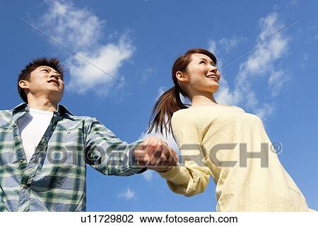 Stock Image - Low angle view of young couple walking and holding hands against a blue sky. Fotosearch