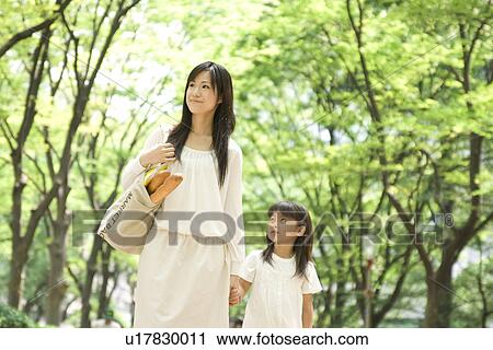 Stock Image - Mother carrying grocery bag and daughter walking through park. Fotosearch