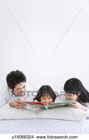 Picture - Mother, father and daughter lying on bed reading picture book. Fotosearch