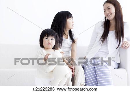 Stock Image - One year old girl holding teddy with mother and sister in the background. Fotosearch