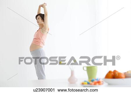 Stock Image - Pregnant woman standing stretching in dining room. Fotosearch