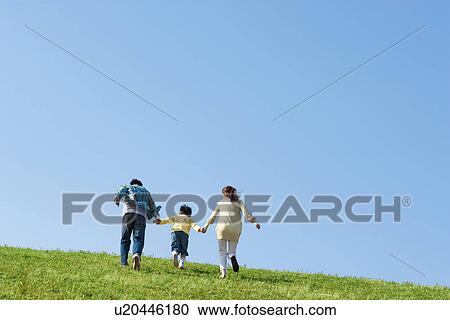 Stock Image - Rear view of a family with young son running up a hill at the park against a blue sky. Fotosearch