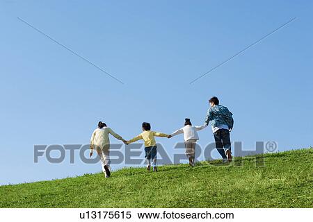 Stock Photography - Rear view of family of four walking and holding hands at the park against a blue sky. Fotosearch