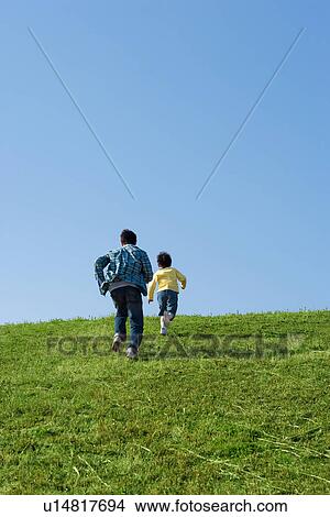 Picture - Rear view of father and son running up a hill against a clear blue sky. Fotosearch