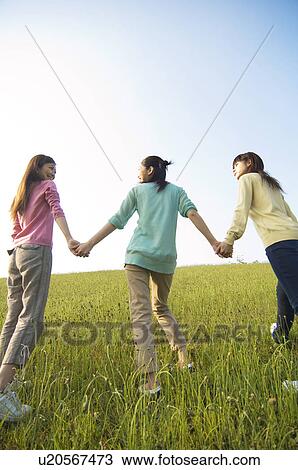 Stock Image - Three female friends walking hand in hand through a field. Fotosearch