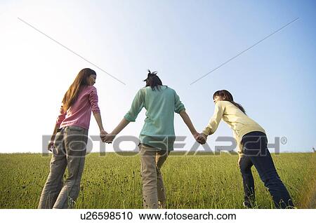 Stock Image - Three female friends walking hand in hand through a field. Fotosearch