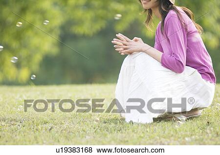 Stock Photo - Woman crouching in a field with bubbles floating around her. Fotosearch