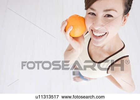 Stock Photo - Woman holding an orange and weighing herself on bathroom scales. Fotosearch