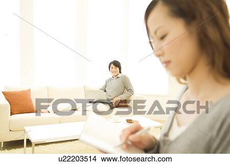 Woman writing in her diary while man in background uses laptop View Large Photo Image Picture - Woman writing in her diary while man in background uses laptop. Fotosearch