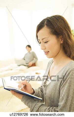 Woman writing in her diary while man in background uses laptop View Large Photo Image Stock Photograph - Woman writing in her diary while man in background uses laptop. Fotosearch