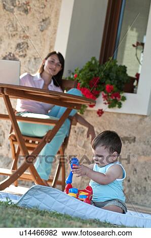 Stock Image - Baby playing with toy and young woman at laptop computer. Fotosearch