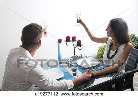 Stock Image - Couple sitting at table toasting with white wine and holding hands. Fotosearch