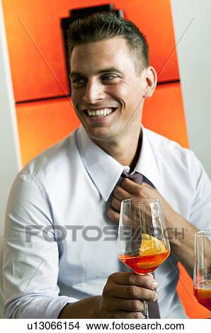 Picture - Young man in business attire with glass of orange wine adjusting tie. Fotosearch