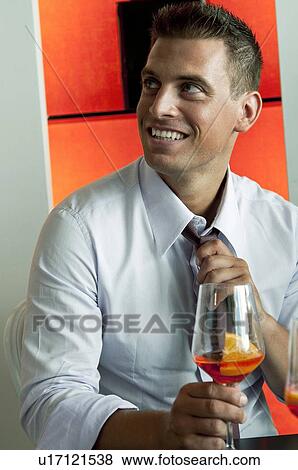 Stock Photo - Young man in business attire with glass of orange wine adjusting tie. Fotosearch