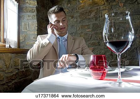 Picture - Young man in suit with mobile phone at a restaurant checking time. Fotosearch