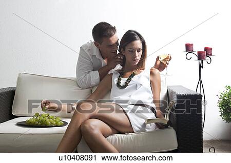 Stock Image - Young man leaning on couch with white wine whispering in young woman's ear reading book and eating grapes. Fotosearch